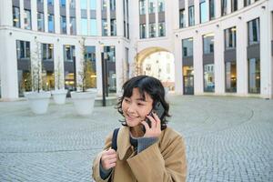 celular tecnologia. jovem coreano mulher fala em Móvel telefone, faz uma telefone ligar em dela caminho lar, anda em baixa rua, cidade Centro, tem Telefone conversação foto