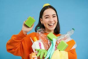 retrato do entusiasmado sorridente mulher mostrando limpeza esponja, esvaziar plástico garrafas e lixo, reciclando, em pé sobre azul fundo foto