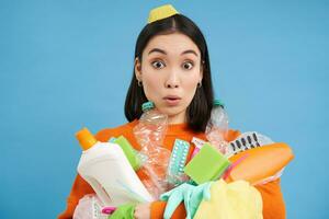 retrato do ásia mulher com surpreso face, detém plástico esvaziar garrafas e lixo para reciclando, Ordenação estações, azul fundo foto