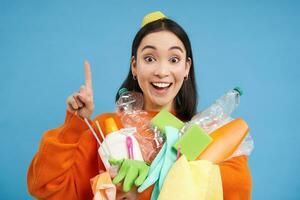 feliz jovem mulher apontando acima, segurando plástico esvaziar garrafas e lixo para reciclando, mostrando eco bandeira, Ordenação estação anúncio, azul fundo foto