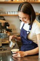 sorridente ásia menina barista, cafeteria funcionários derramando cozido no vapor leite dentro café, preparar cappuccino com café com leite arte, em pé dentro azul avental atrás contador foto