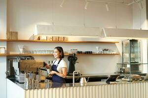 retrato do cafeteria contador com barista menina trabalhando com café máquina, fazer ordem para cliente dentro comprar, vestindo azul avental foto