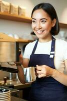 vertical tiro do barista, ásia menina vapor leite para capuccino, preparar café com leite para cliente, vestindo azul avental, sorridente Felizmente, trabalhando dentro cafeteria foto