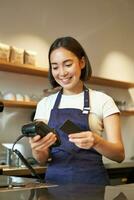 retrato do sorridente ásia barista, café fazer compras empregado usando pos terminal e crédito cartão, ajuda cliente pagar sem contato dentro cafeteria foto