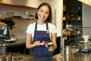 retrato do ásia barista menina às contador, mostrando cartão máquina para cliente quem quer para pagar sem contato, levando ordem, em pé dentro cafeteria foto