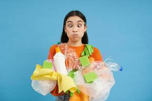 jovem mulher detém plástico lixo, reciclável lixo e estrabismo dela olhos, confuso de reciclando regras, azul fundo foto