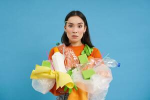 confuso ásia menina não conhecer quão para reciclar. mulher detém plástico lixo com chocado face, azul fundo foto