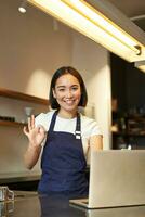 retrato do barista garota, ásia fêmea cafeteria trabalhador, mostra OK, Está bem placa dentro aprovação, recomendar smth, feliz e satisfeito, vestindo avental, em pé com computador portátil atrás contador foto