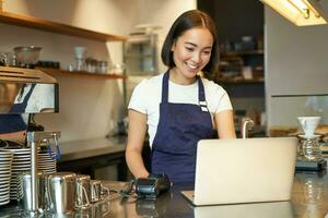 retrato do jovem menina cafeteria proprietário, olhando às dela computador portátil, levando ordem, servindo cliente dentro café fazer compras foto