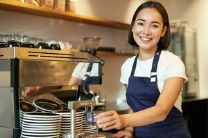 retrato do fofa barista menina trabalhando atrás contador, fazer café, vapor leite para capuccino, vestindo cafeteria uniforme azul avental foto