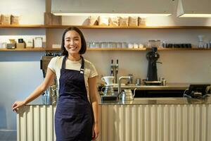 retrato do sorridente ásia fêmea barista, vestindo avental, em pé perto contador com café, trabalhando dentro cafeteria foto