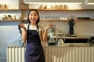 entusiasmado jovem ásia mulher, barista dentro avental, olhando espantado, aplaudir mãos e sorridente, em pé dentro café comprar, trabalhando dentro cafeteria foto