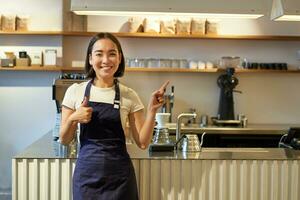 feliz jovem ásia barista, fêmea funcionários trabalhador dentro avental, mostrando polegares acima e apontando às cópia de espaço, demonstrando cafeteria propaganda foto