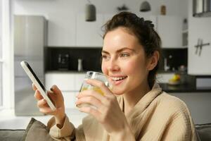 fechar acima retrato do rindo, sorridente jovem mulher goza manhã, bebendo laranja suco e segurando Smartphone, lendo notícia em Móvel aplicativo foto