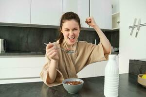 retrato do entusiasmado jovem mulher comendo cereais com leite, olhando animado e feliz, sentado perto cozinha bancada e tendo café da manhã, levantando mão acima dentro triunfo foto