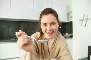 retrato do feliz jovem mulher inclina-se em cozinha bancada e comendo cereais, tem leite e tigela dentro frente do dela, tendo dela café da manhã, vestindo roupão de banho foto