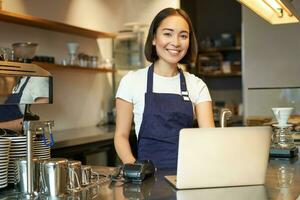 retrato do jovem menina cafeteria proprietário, olhando às dela computador portátil, levando ordem, servindo cliente dentro café fazer compras foto