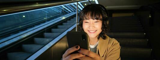 retrato do feliz ásia menina dentro fones de ouvido, sorridente e olhando às Smartphone, assistindo vídeo ou ouvindo música em Móvel telefone, sentado em Escadaria foto