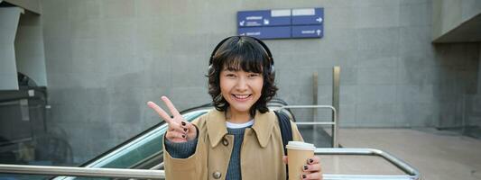 positivo coreano menina dentro fones de ouvido, bebidas café para ir, mostra paz, sinal v, carrinhos em rua e sorrisos alegremente foto