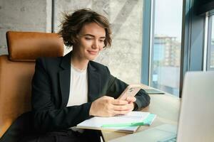 retrato do morena vendedora, corporativo mulher dentro terno, trabalhando dentro escritório, sentado com Smartphone e computador portátil, respondendo em mensagem foto