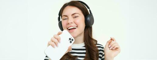 tecnologia e social meios de comunicação. retrato do feliz jovem mulher cantando karaokê, jogando música aplicativo em Smartphone, vestindo sem fio fones de ouvido, em pé sobre branco fundo foto