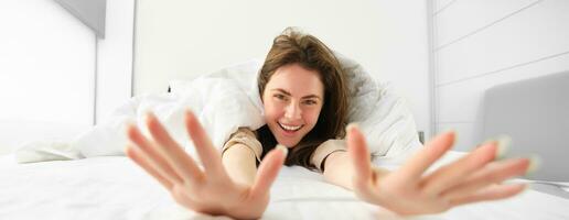 lindo mulher alongamento, mostrando dela braços para dentro Câmera, deitado dentro cama, acordando acima dentro manhã a partir de Boa dormir ou sesta foto