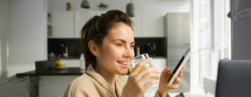 retrato do boa aparência mulher às lar, usando Móvel telefone e bebendo manhã vidro do laranja suco foto