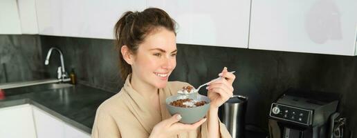 fechar acima retrato do sorridente, lindo mulher comendo cereais com leite, segurando colher e tigela, olhando lado de fora janela dentro manhã, em pé perto cozinha bancada foto