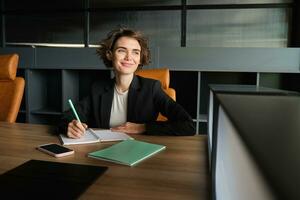 retrato do bem sucedido vendedora, corporativo mulher dentro terno, sentado dentro cadeira dentro dela escritório com documentos, escrevendo baixa notas para encontro apresentação, fazendo dela trabalho, olhando lado de fora janela foto