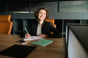 retrato do bem sucedido morena mulher dentro terno, corporativo Gerente dentro escritório, escrita, preparando documentos para reunião, lendo caso foto