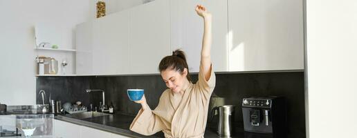 retrato do feliz menina dançando com café dentro a cozinha, vestindo roupão de banho, desfrutando dela manhã rotina foto