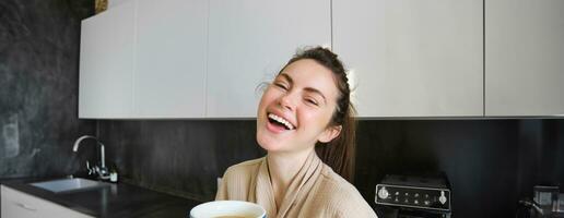 retrato do boa aparência jovem mulher, bebendo café dentro a cozinha, desfrutando dela manhã rotina e sorridente às a Câmera foto