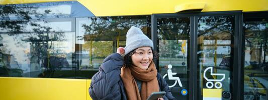 retrato do menina em pé perto ônibus em uma parar, esperando para dela público transporte, cheques cronograma em Smartphone aplicativo, detém Móvel telefone, desgasta caloroso roupas foto