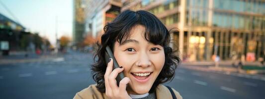 retrato do feliz coreano garota, fala em Móvel telefone, parece surpreso e feliz, recebe positivo ótimo notícia sobre Telefone conversação, carrinhos em rua do cidade foto