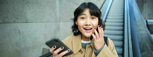 alegre e positivo coreano garota, comemora, parece surpreso, vai baixa escada rolante com Smartphone e parece espantado de smth foto