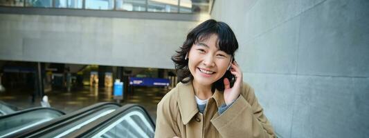 retrato do fofa sorridente coreano garota, vai acima escada rolante, escuta música dentro sem fio fones de ouvido e usa Móvel telefone, detém Smartphone foto