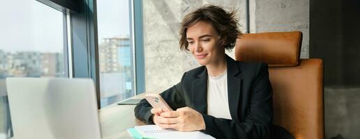 retrato do morena vendedora, corporativo mulher dentro terno, trabalhando dentro escritório, sentado com Smartphone e computador portátil, respondendo em mensagem foto