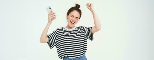 menina dançando com Móvel telefone contra branco fundo. mulher com Smartphone cantando, levantando mãos acima e a comemorar foto