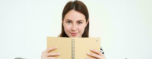 imagem do jovem mulher com caderno, segurando dela pessoal diário, trabalhos planejador, sorridente, lendo algo, em pé sobre branco fundo foto