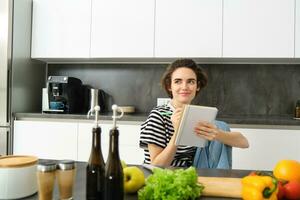 retrato do jovem mulher olhando às cozinhando ingredientes em cozinha contador e fazer notas, escrevendo baixa receitas, pensando do refeição para jantar, preparando vegetariano Comida foto