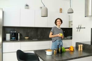 retrato do feliz, saudável jovem mulher, vegetariano fazer ela mesma salada, posando perto legumes em cozinha, cortar ingredientes para vegano refeição foto