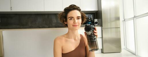 ginástica instrutor, sorridente jovem mulher exercite-se às lar, senta em ioga esteira e bebidas água, faz exercícios, Treinamento sessão dentro dela cozinha foto