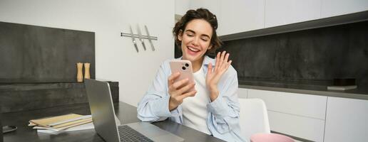 retrato do jovem mulher vídeo chats com amigo em Smartphone, ondas mão às Móvel telefone Câmera, chamadas alguém usando inscrição foto