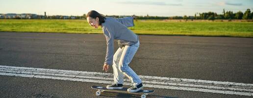 feliz skatista menina equitação dela skate e tendo Diversão em esvaziar rua. sorridente mulher desfrutando cruzador passeio em ensolarado estrada foto