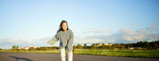 fofa ásia menina equitação skate, patinação em estrada e sorridente. skatista em cruzador Longboard desfrutando ao ar livre em ensolarado dia foto