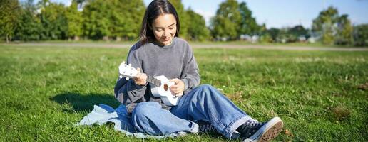 despreocupado ásia menina cantando e jogando ukulele dentro parque, sentado em grama, músico relaxante em dela livre Tempo ao ar livre foto