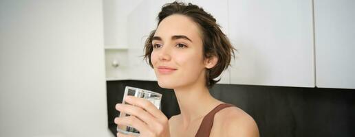 fechar acima retrato do sorridente ginástica mulher, segurando vidro do água e olhando longe satisfeito. menina bebidas depois de exercite-se foto