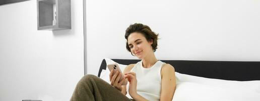 retrato do jovem lindo mulher deitado dentro cama, em repouso dentro quarto, Mensagens, usando Móvel telefone, segurando Smartphone e sorridente, rolagem social meios de comunicação, gastos conforto Tempo às casa foto