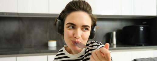 fechar acima retrato do jovem sorridente mulher dentro fones de ouvido, comendo cereais com colher e ouvindo música, vestindo fones de ouvido, sentado dentro a cozinha foto