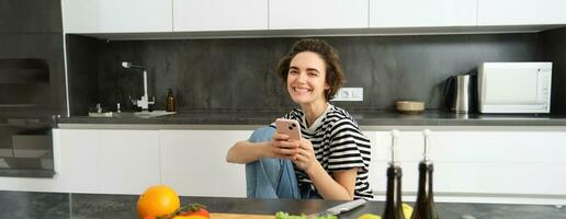 retrato do jovem mulher procurando para cozinhando receitas conectados usando Smartphone, sentado perto vegetais, salada ingredientes e cortar quadro, sorridente às Câmera foto